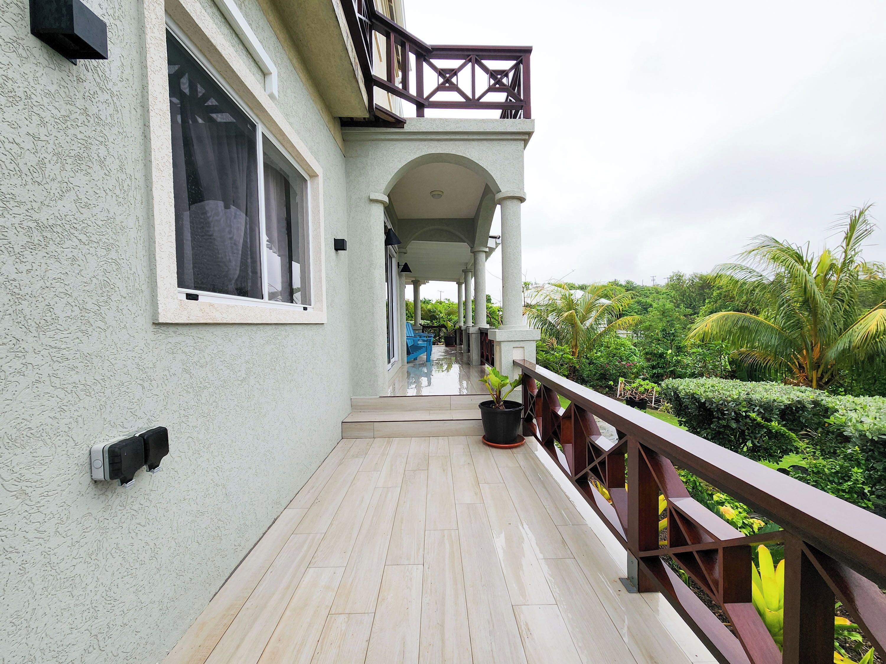 A covered outdoor area with a wooden deck, a railing, and a view of lush greenery.
