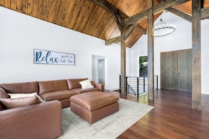 A spacious living room with a brown leather sectional sofa, a matching ottoman, a wooden ceiling, and a chandelier.