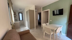 Ein Wohnzimmer mit einer Couch, einem Esstisch mit Stühlen, einem an der Wand montierten Fernseher und einer Küche im Hintergrund.
