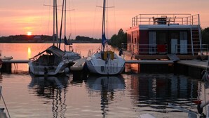 A marina at sunset with boats docked, a small building with a balcony, and calm water reflecting the sky. - Hausboot in Barth am Barther Bodden (Barth)