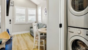 A laundry room with stacked washers and dryers, a gray sofa, a wooden dining table, and a white door with a window. - Bayside Bliss (Lower Township)