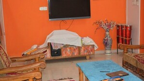 A living room with a television mounted on an orange wall, a wooden sofa, a blue coffee table, and a decorative vase with flowers. - Charming 3-bedroom bungalow with AC, fitness room in wonderful Gwalior (GWALIOR)