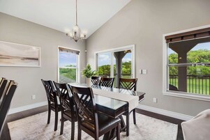 Un comedor con mesa de vidrio, sillas de madera oscura y una araña. Hay ventanas amplias con vistas a zonas verdes y una pérgola.