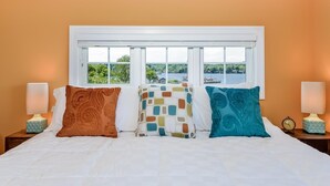 Una cama con tres almohadas, una ventana con vistas al agua y vegetación, y dos mesitas de noche con lámparas y relojes.