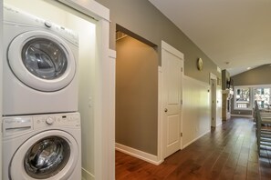 A laundry room with two front-loading washers and dryers, a door leading to another room, and a hallway with wooden flooring and a clock on the wall. - Four Generations (Saugatuck)