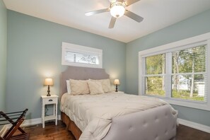 A bedroom with a bed, two bedside tables with lamps, a ceiling fan, a window with blinds, and a view of trees.