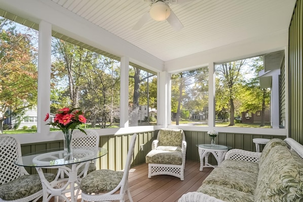 A covered porch with wicker furniture, a glass table, and a view of a residential neighborhood.