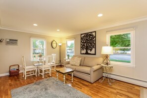 A living room with a beige sofa, white chairs, a coffee table, and a gray rug. There are two windows with views of a parking lot and trees. A wall-mounted clock and a lamp are visible. - Water Street Retreat (Saugatuck)