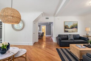 Uma sala de estar moderna com um sofá de couro escuro, uma mesa de centro redonda e uma luminária pendente de vime. Um quadro está pendurado na parede, e uma área de jantar é visível ao fundo.