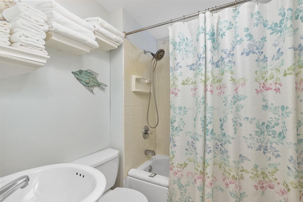 A bathroom with a white sink, toilet, and shower. There are towels stacked on a shelf and a shower curtain with a floral pattern.