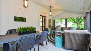 A covered patio area with a dining table, chairs, and a sofa. There are hanging lanterns, potted plants, and a view of the outside.