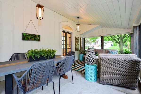 A covered patio area with a dining table, chairs, and a sofa. There are hanging lanterns, potted plants, and a view of the outside. - Saugatuck Farmhouse (Saugatuck)