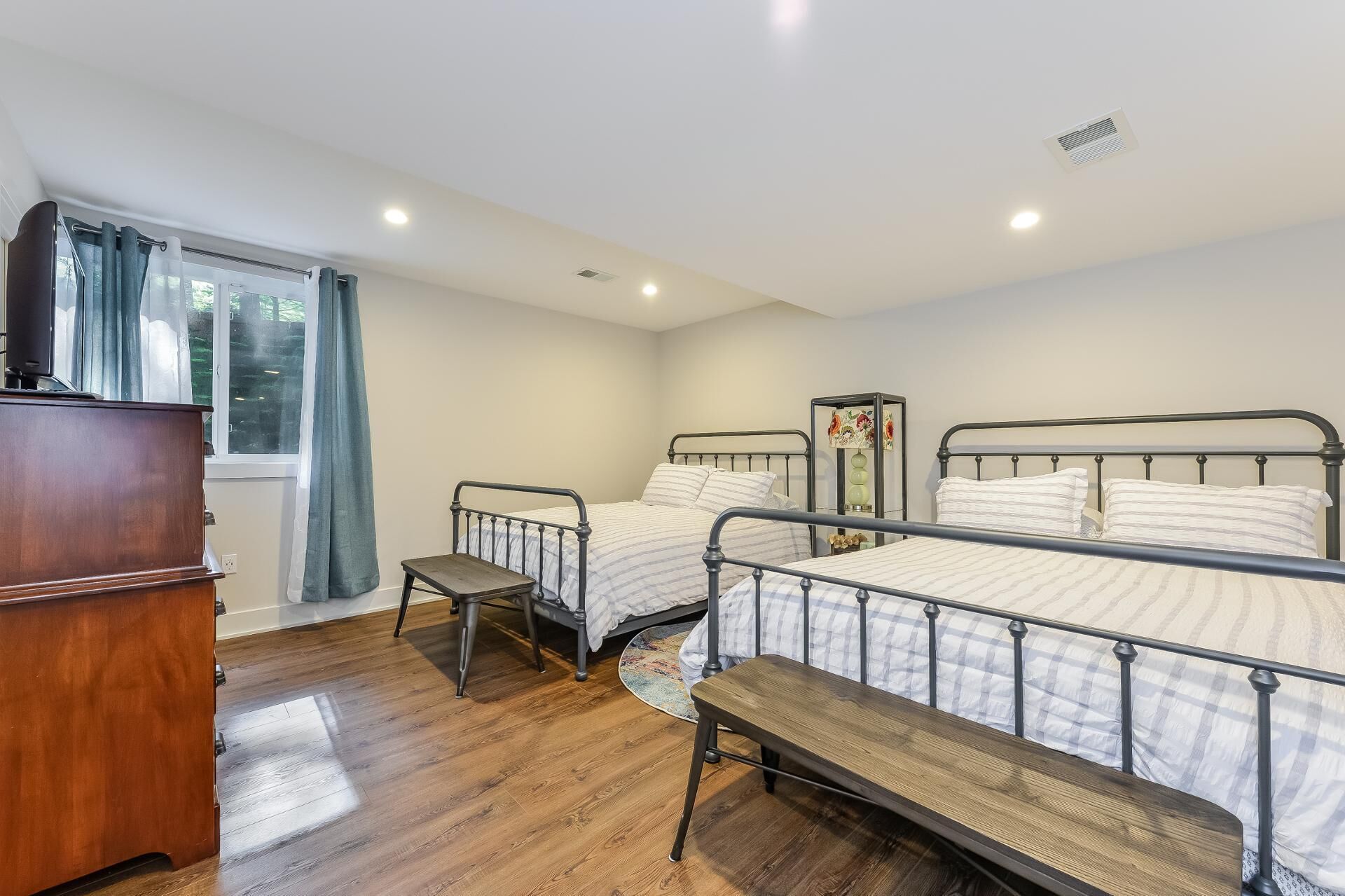 A bedroom with two metal-framed beds, a wooden bench, a dresser, and a window with curtains.