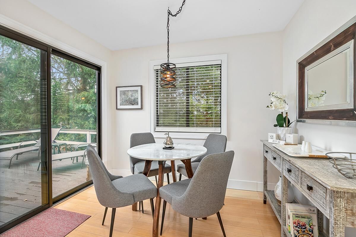 A modern dining area with a round table, grey upholstered chairs, a hanging light fixture, and a view through large windows.