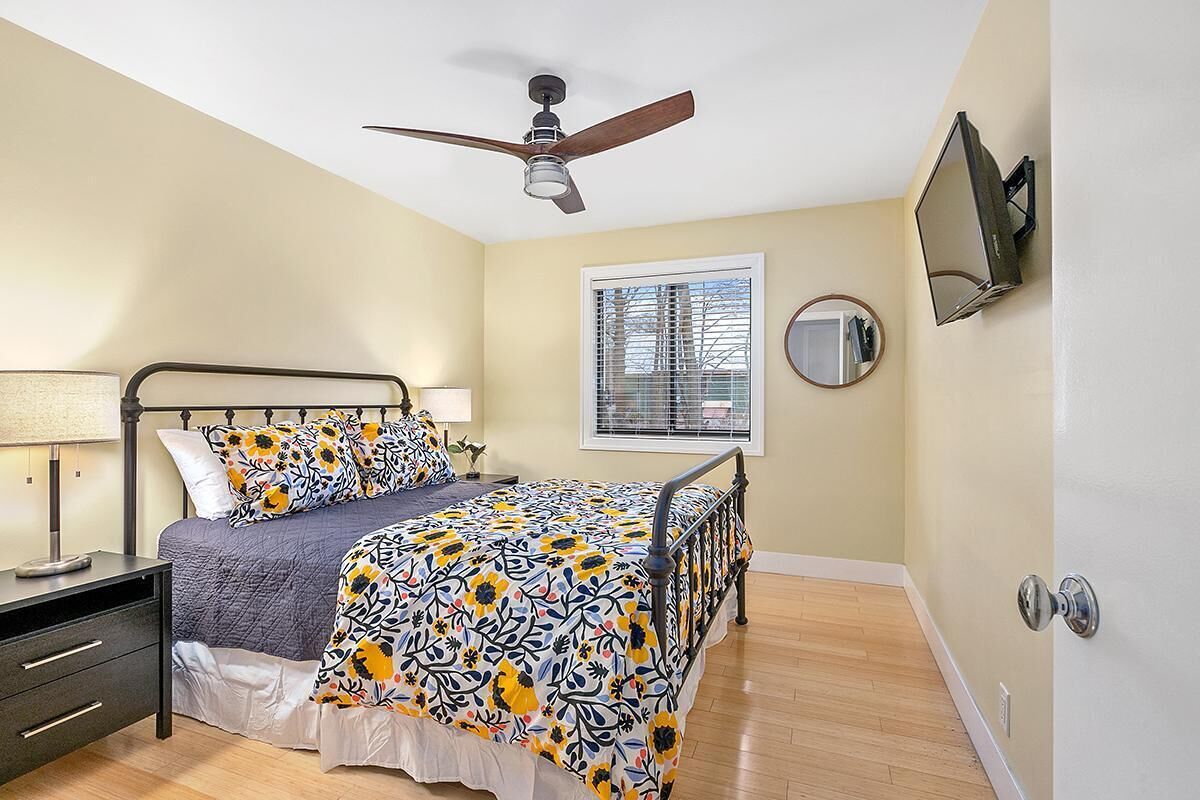 A bedroom with a bed, bedside tables, a ceiling fan, a window with blinds, a wall-mounted TV, and a door with a handle.