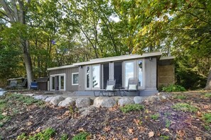 Una cabaña moderna con grandes puertas correderas de vidrio, un patio con mobiliario exterior y un muro de contención de piedra.
