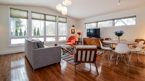 Un salon spacieux avec un canapé gris, une table à manger en bois et des chaises, ainsi qu’un téléviseur. De grandes fenêtres laissent entrer la lumière naturelle et offrent une vue sur les arbres à l’extérieur.