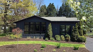 Une maison moderne à deux étages, dotée d’une façade sombre, de grandes fenêtres et d’un jardin bien entretenu avec divers arbustes et une pelouse.