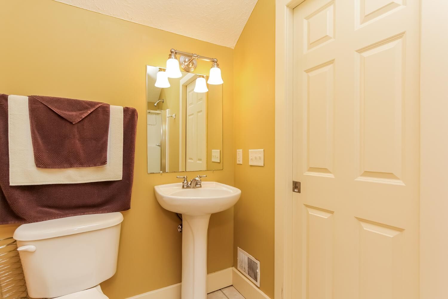 A bathroom with a white toilet, a pedestal sink, and a mirror with three lights.