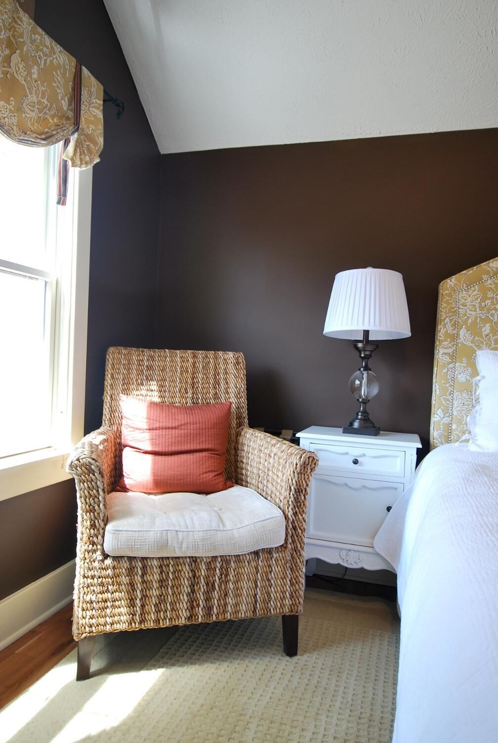 A cozy bedroom with a wicker chair, a bedside table with a lamp, and a bed with a patterned headboard.