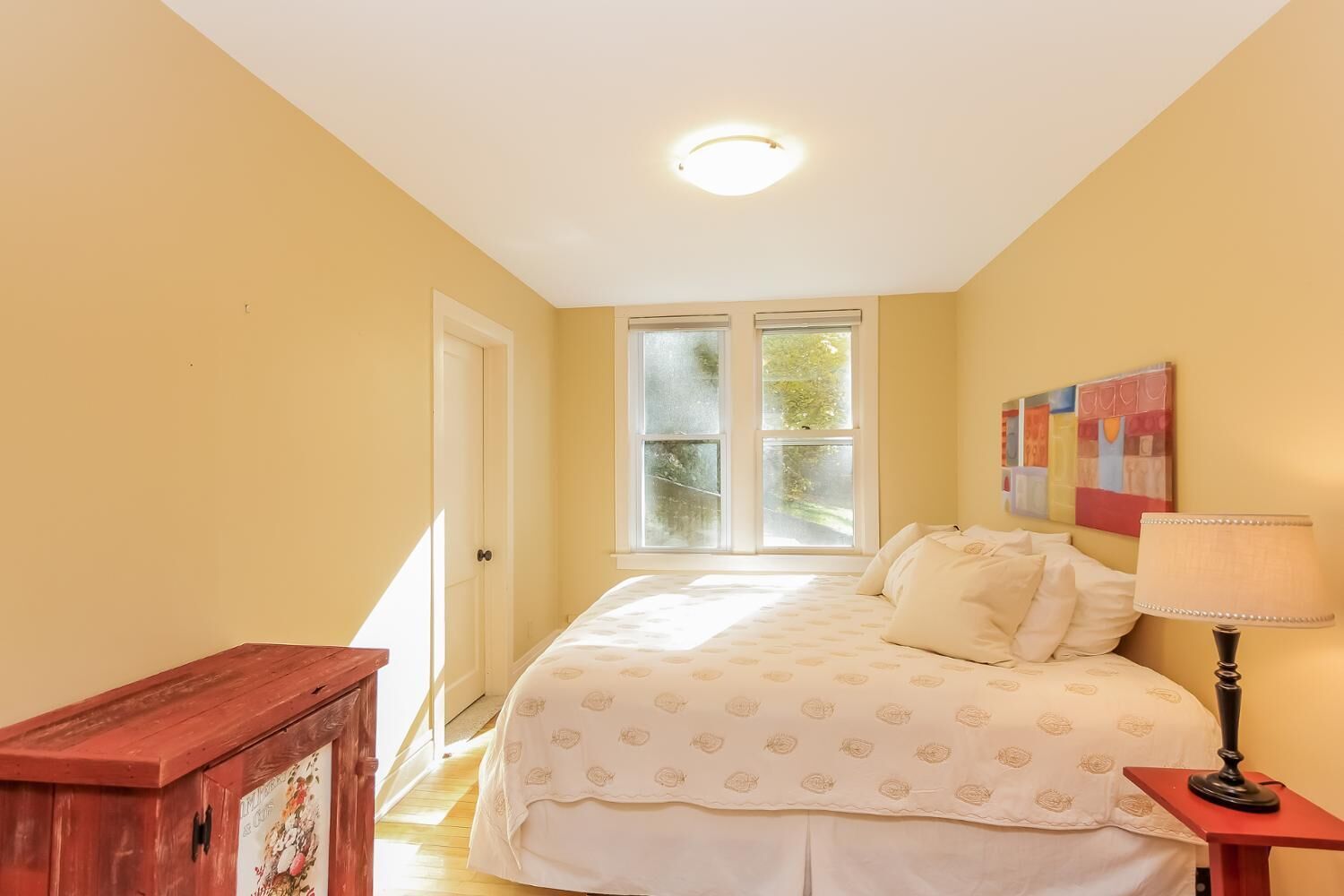 A bedroom with a bed, a nightstand, a wooden cabinet, a window with blinds, and a ceiling light.