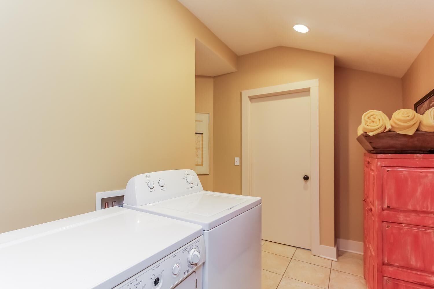 A laundry room with a front-loading washer and dryer, a wooden cabinet with folded towels, and a door leading to another room.