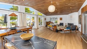 A spacious living area with a wooden ceiling, large windows, a dining table with a bowl, and a fireplace. - How you Dune (Saugatuck)
