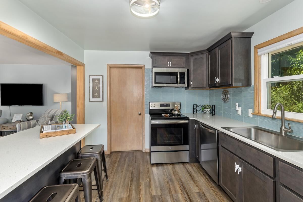 A modern kitchen with dark cabinets, a stainless steel oven, a microwave, and a large window providing a view of greenery.