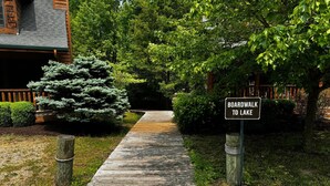 A wooden boardwalk leading to a lake, surrounded by lush greenery and a wooden cabin.
