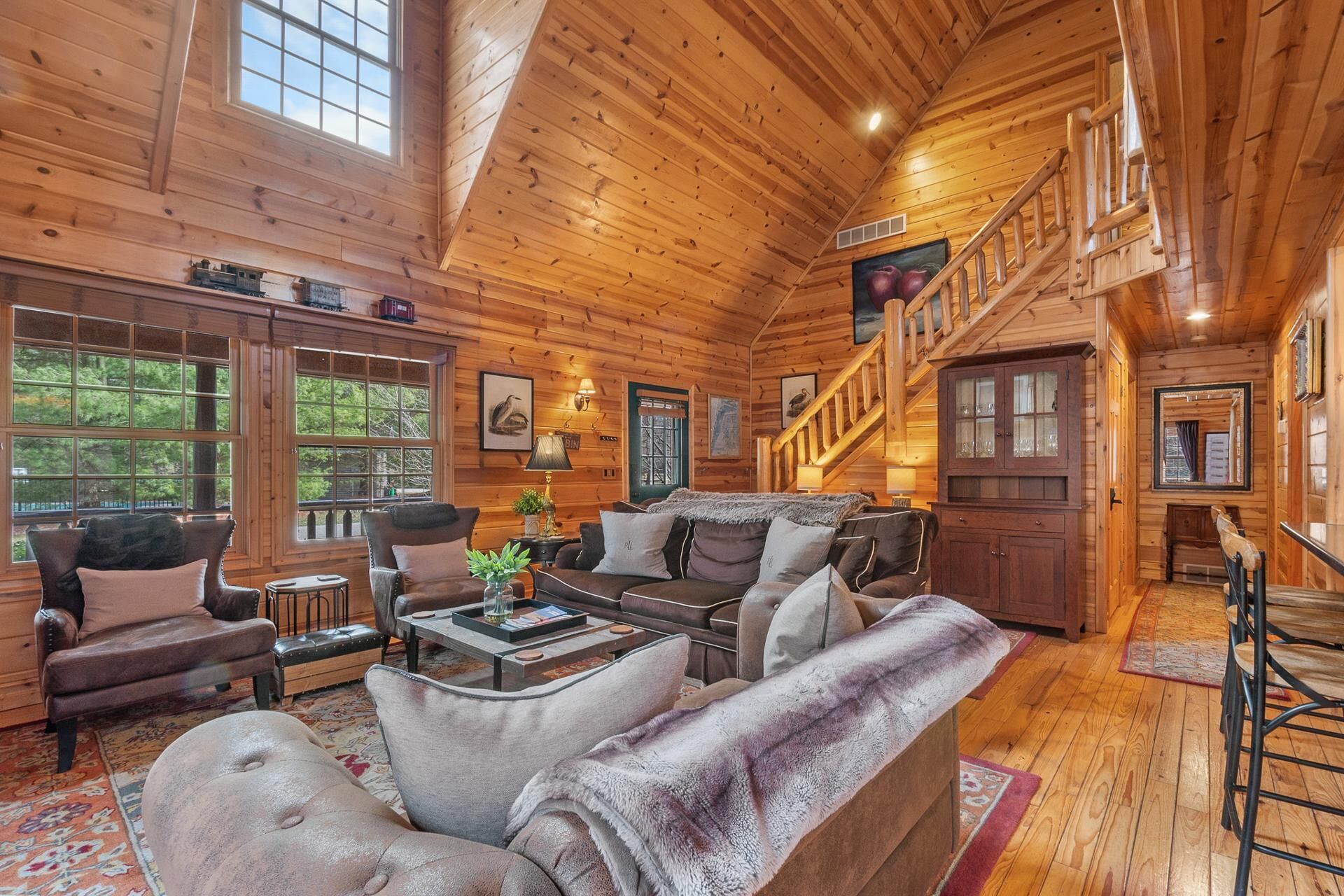 A cozy log cabin interior with a living room, a staircase, and large windows.