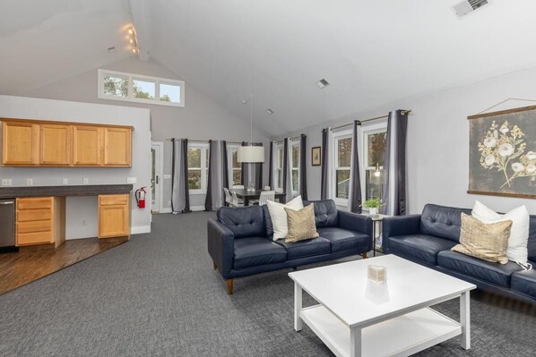 A spacious living area with a dark blue sofa set, a white coffee table, and a kitchen area with wooden cabinets and a stainless steel refrigerator.