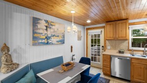 A modern kitchen with wooden cabinets, a stainless steel dishwasher, and a dining area with a blue sofa and table. - "cozy Cottages" Green Cottage Hot Tub-town! (Saugatuck)