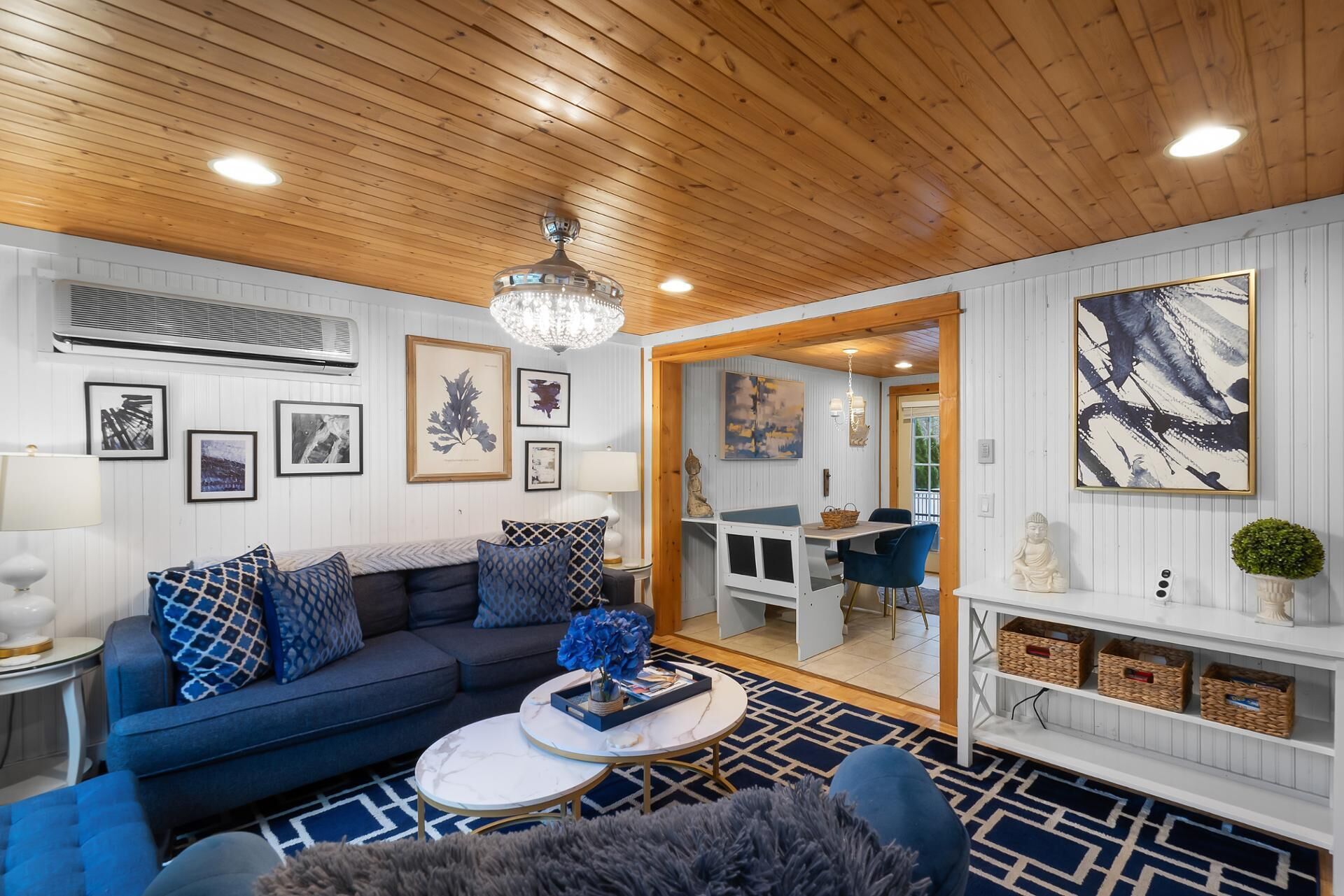 A modern living room with a wooden ceiling, a blue sofa, a white coffee table, and a dining area with a blue chair.