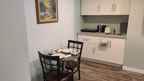 Un coin repas avec une table dressée pour deux personnes, un tableau accroché au mur et une petite cuisine équipée de placards et d’appareils électroménagers.