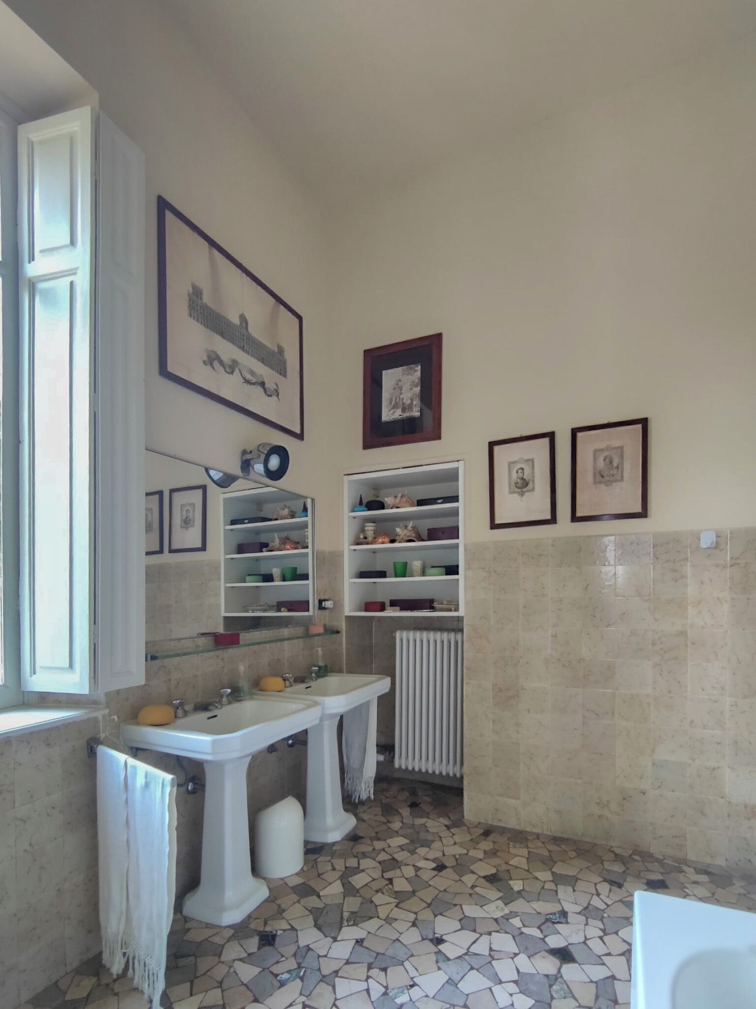 A bathroom with two sinks, a mirror, a radiator, and shelves with decorative items.