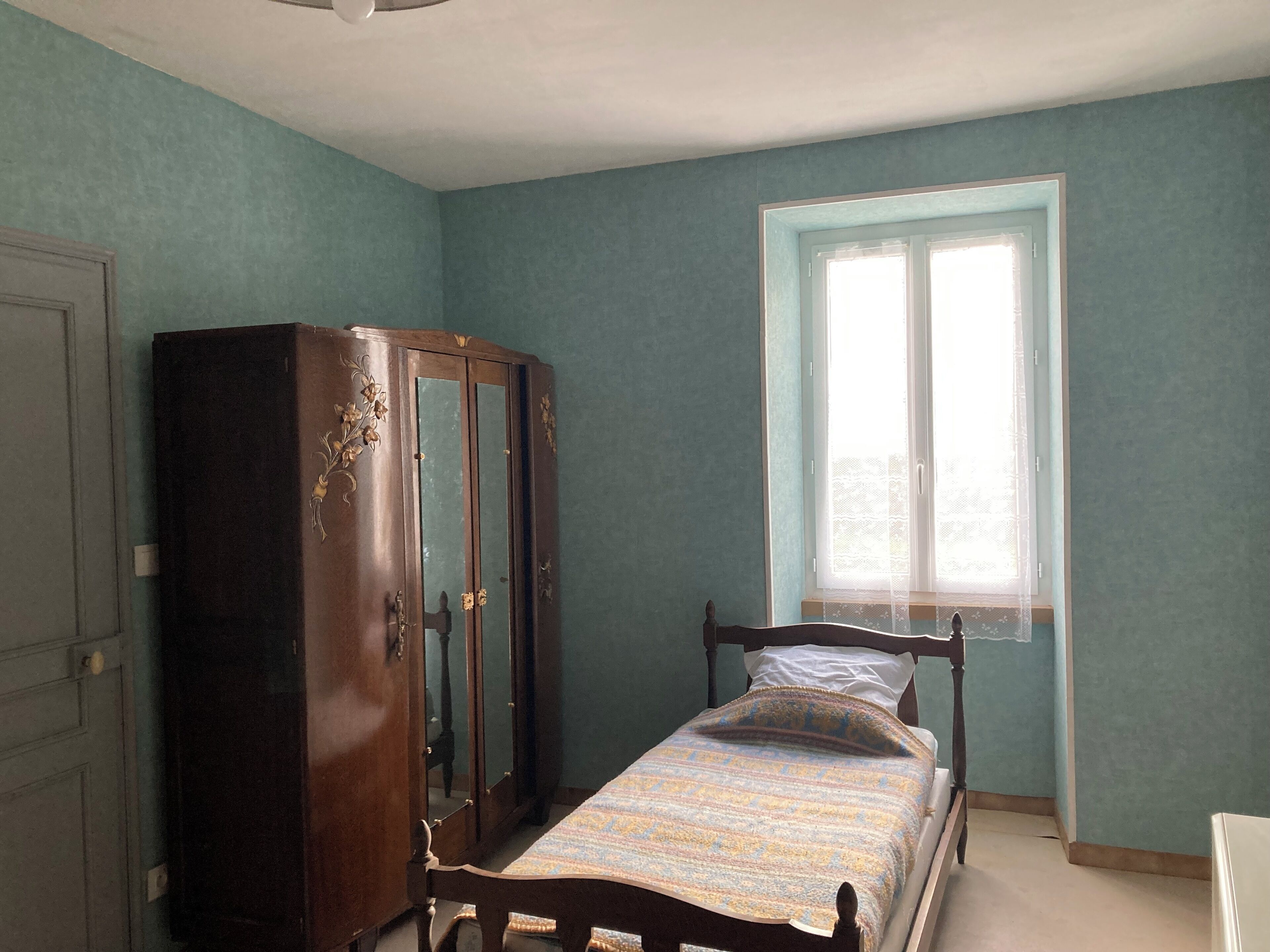 A room with teal walls, a wooden bed with a patterned bedspread, a wooden wardrobe, and a window with sheer curtains.
