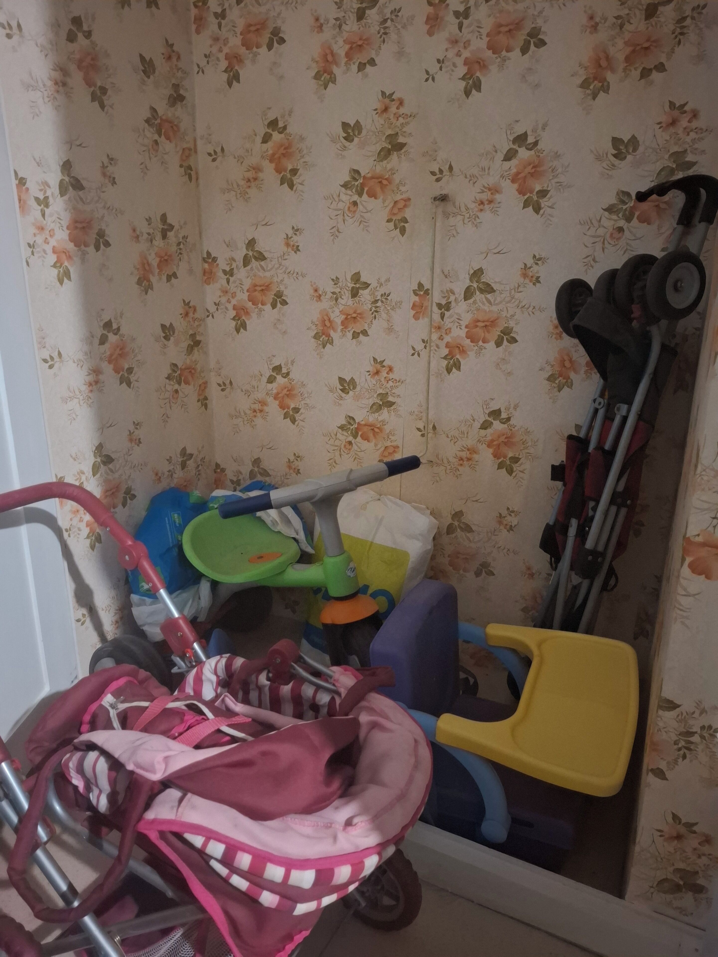 Een kamer met bloemenbehang, een groen en blauw speelautootje, een gele stoel en een kinderwagen.