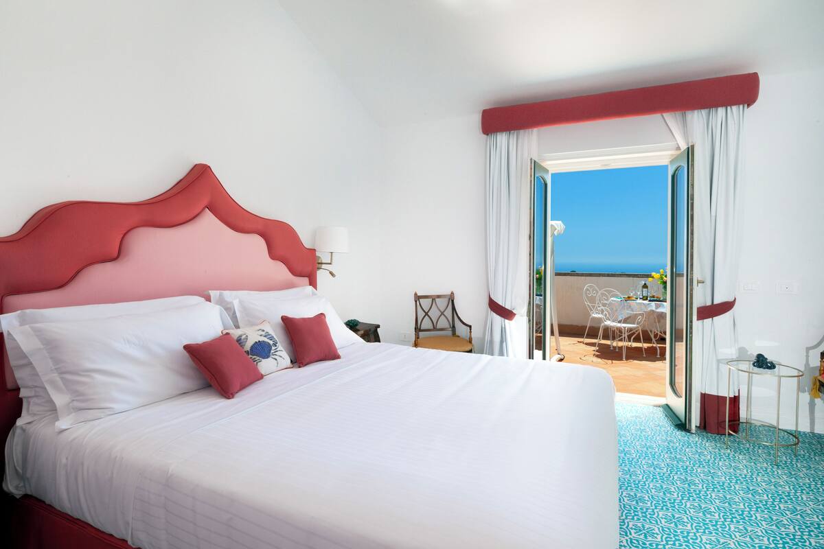 A hotel room with a large bed, a view of the sea, and a balcony with outdoor seating.