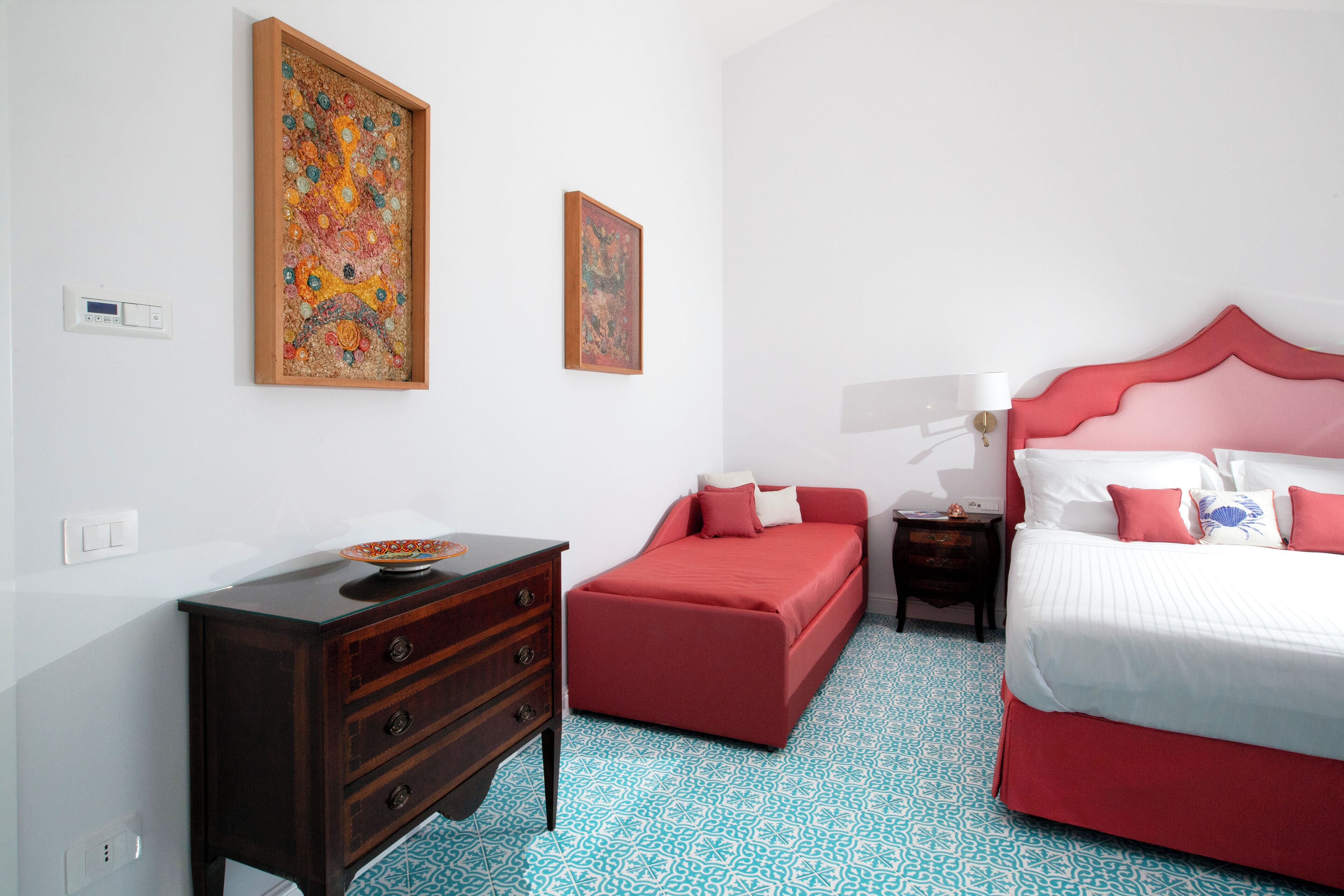 A bedroom with a red bed, a wooden dresser, a red ottoman, and two framed artworks on the wall.