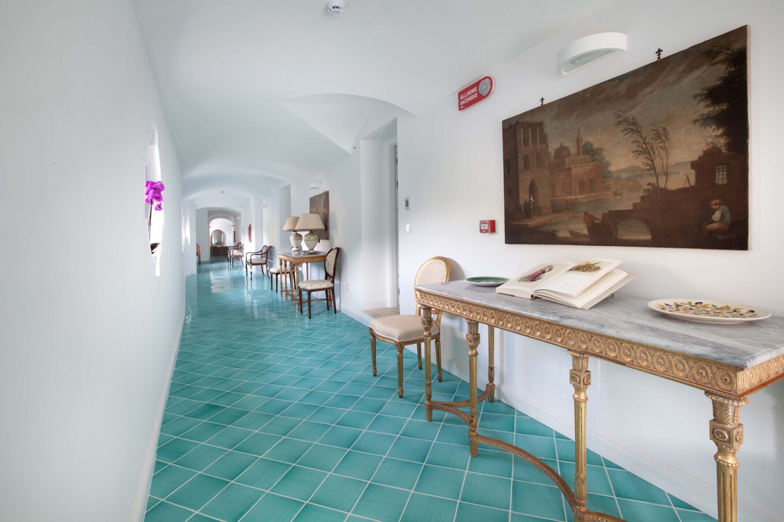 Un pasillo de hotel con piso de baldosas azules, una mesa de comedor con sillas y un cuadro grande en la pared.