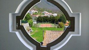 A view through a window with a decorative frame, revealing a tennis court, a green area, and buildings in the background. - De Pando Panoramic Suites 1086 (Scala)