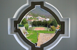 Vista a través de una ventana con un marco decorativo, que muestra una cancha de tenis, una zona verde y edificios al fondo.