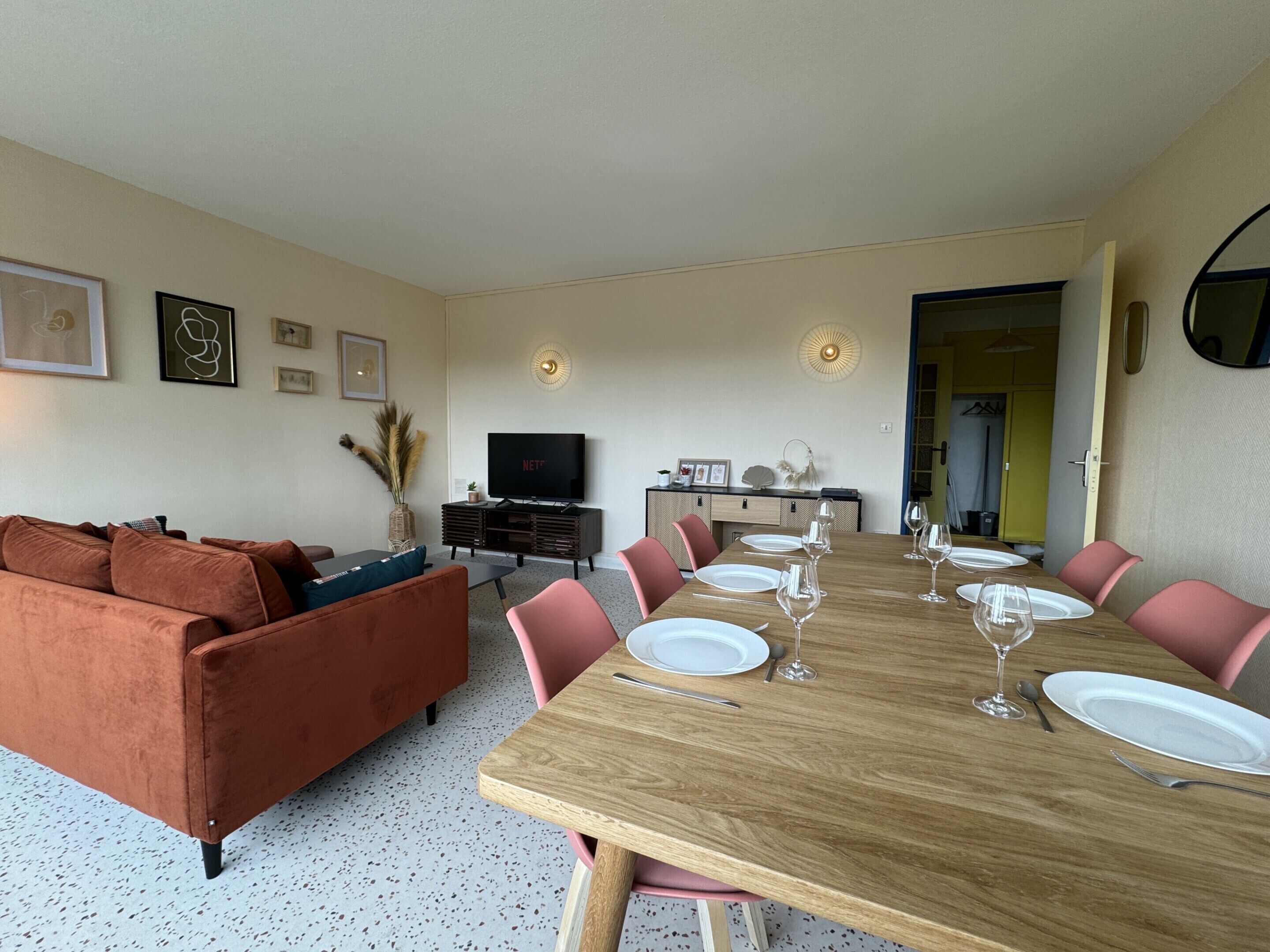 Een moderne woonkamer met een houten eettafel gedekt voor zes personen, een bruine bank en een televisie op een zwart tv-meubel.