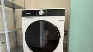 A white washing machine with a black door, labeled "VALBERG" and "INVERTER TECHNOLOGY," placed on a tiled floor next to a metal shelf and a teal cabinet. - Apartment in Le Mans Near 24H Circuit (Le Mans)