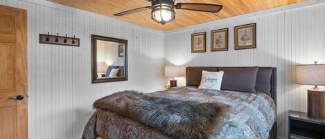 Une chambre avec un plafond en bois, un lit recouvert d’un plaid en fourrure, un ventilateur de plafond, un miroir et, au mur, des reproductions encadrées de motifs botaniques.