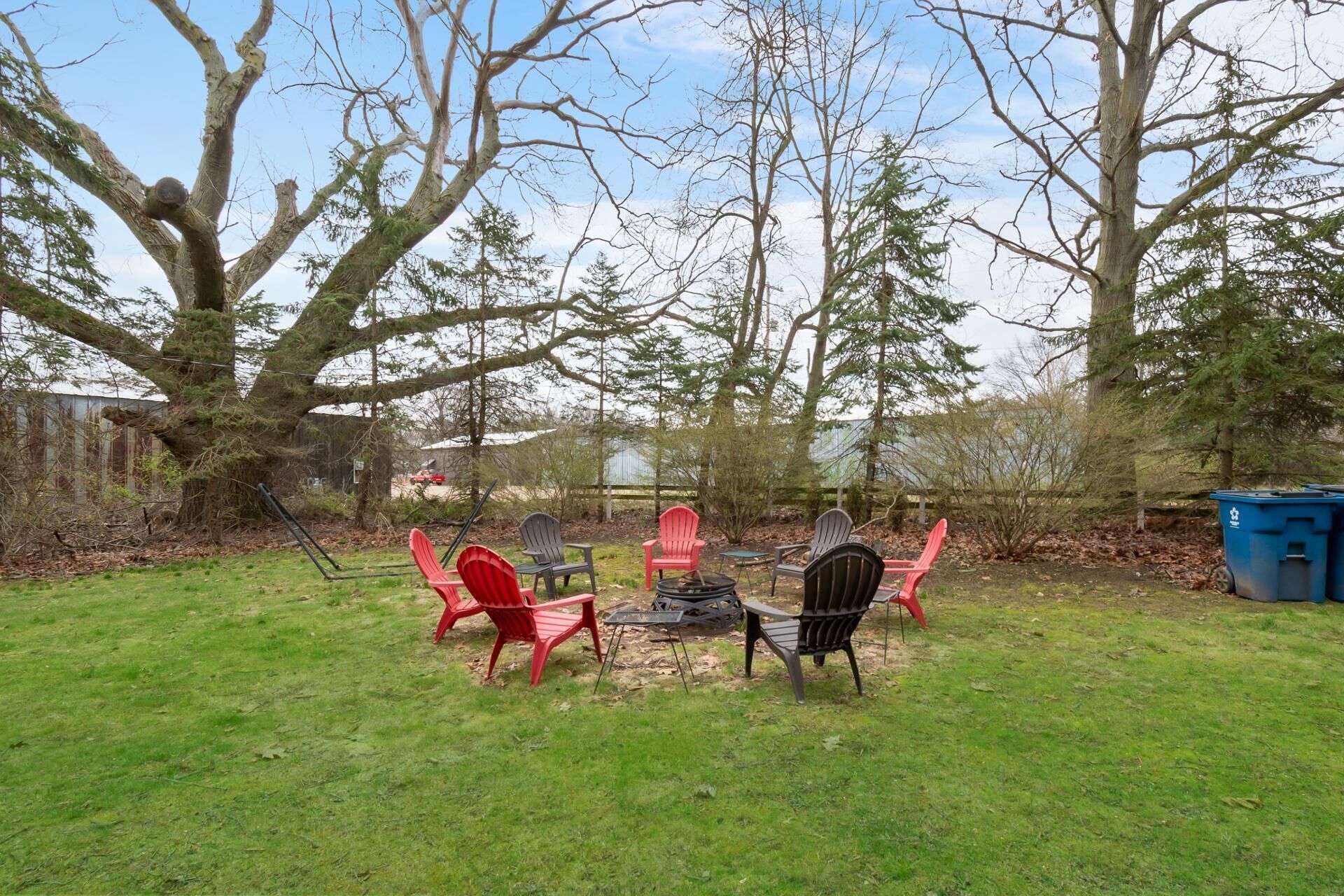 Ein Garten mit Feuerstelle, roten und schwarzen Adirondack-Stühlen und einer blauen Mülltonne.