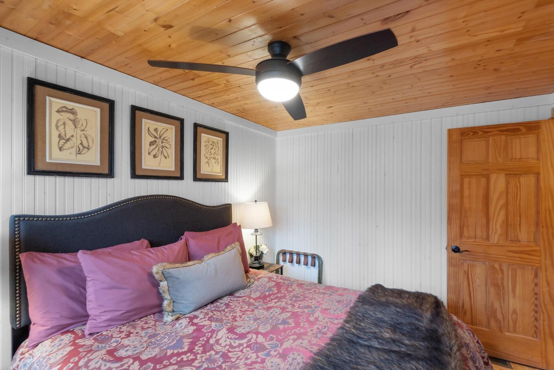 Un dormitorio con techo de madera, cama con ropa de cama estampada, mesita de noche con lámpara y cuadros en la pared.