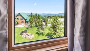 Une fenêtre à cadre en bois donnant sur un chalet en bois entouré d’arbres et bordé d’une zone herbeuse.