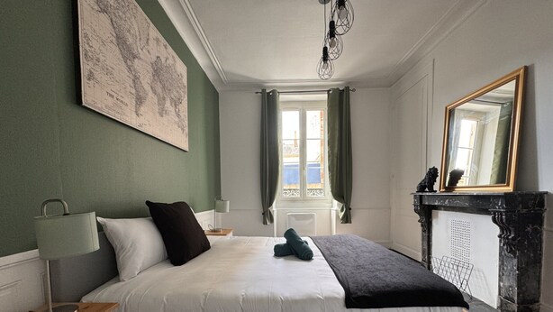 A bedroom with a bed, a nightstand, a lamp, a map on the wall, a window with curtains, and a fireplace. - Refined Apartment With Courtyard in La Mans (Le Mans)