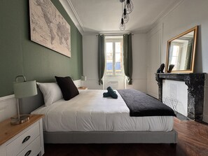 Een slaapkamer met een bed, nachtkastje, lamp, kaart op de muur, raam met gordijnen en een open haard.